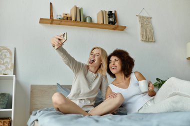 Rahat bir ev elbisesi giymiş iki kadın bir yatakta selfie çekiyorlar, gülüyorlar ve günlerinin tadını çıkarıyorlar..