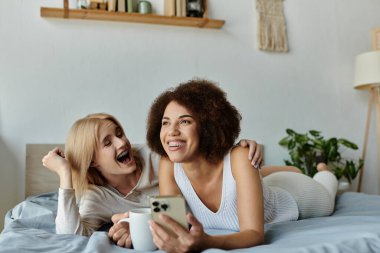 Lezbiyen bir çift yataklarında yatarken birlikte güler, biri bardak tutarken, diğeri de akıllı telefon..