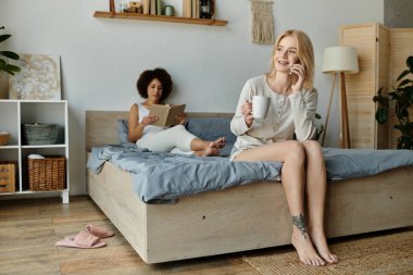 A lesbian couple enjoys a relaxing morning at home, one reading a book while the other sips coffee and talks on the phone. clipart