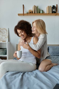 Çok kültürlü lezbiyen bir çift, sıcak bir kupa tutan kıvırcık saçlı bir kadınla yataklarında sevgi dolu bir kucaklaşmayı paylaşıyor..