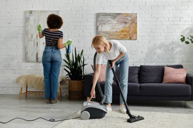 Çok kültürlü lezbiyen bir çift modern dairelerini temizliyor. Biri sanat eserlerini temizlerken diğeri de halıları süpürüyor..