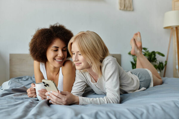Two women in comfortable homewear lie in bed, looking at a smartphone while one enjoys a cup of coffee.