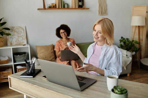 Блондинка за столом в офисе улыбается, пока ее темноволосый партнер сидит на диване с ноутбуком и кофе