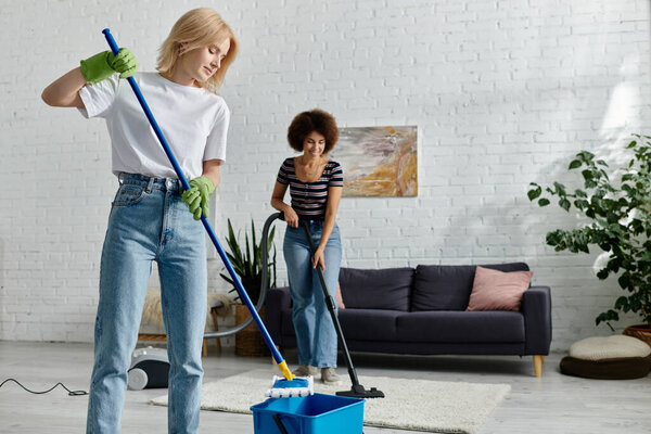 A lesbian couple cleans their modern apartment, one with a mop and bucket, the other with a vacuum.
