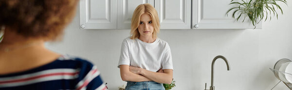 Молодая женщина с светлыми волосами стоит на кухне, скрещенные руки, ее выражение лица смесь недовольства и разочарования.