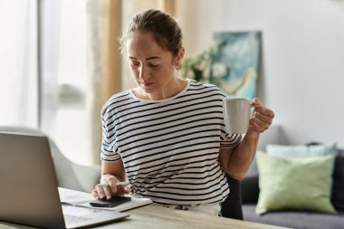 Genç bir kadın evde dizüstü bilgisayarına odaklanırken kahvesini yudumluyor..