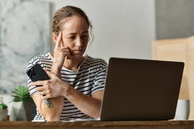 A woman with vitiligo contemplates while using a laptop and holding her phone. clipart