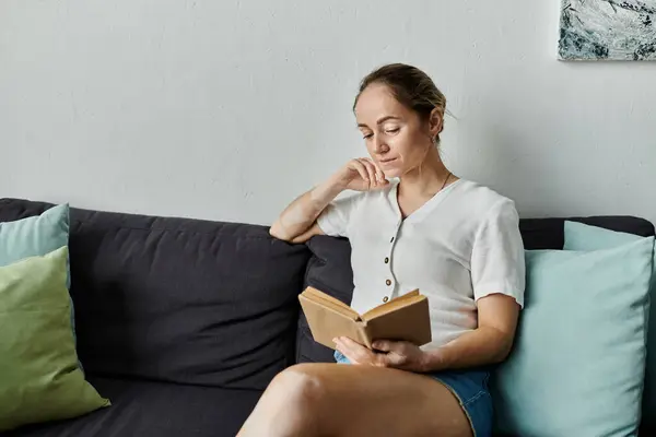 stock image A thoughtful young woman enjoys a book while relaxing on a plush sofa.