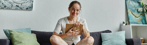 Женщина улыбается, держа книгу, удобно сидя с красочными подушками.