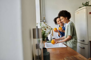 Two women share smiles while washing oranges at their cozy home kitchen. clipart