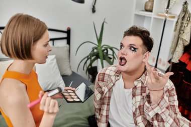 A queer individual joyfully receives a makeup application from their friend.