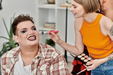 A fashionable individual smiles as their friend applies makeup in a cozy living space. clipart