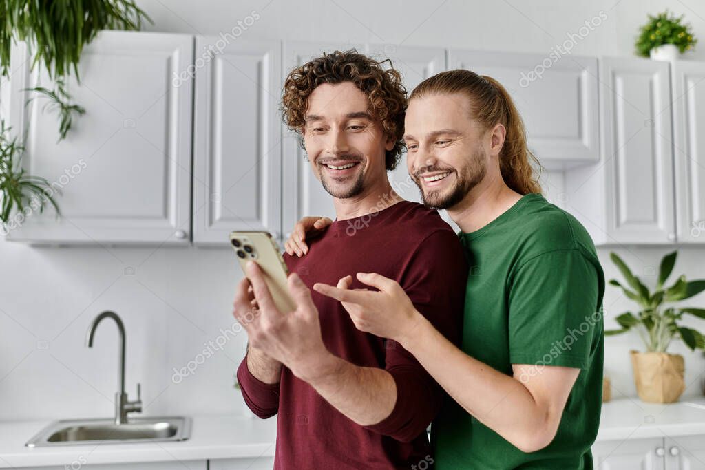 Two men joyfully engage with their smartphone in a modern kitchen.