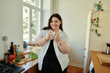 Gülümseyen kadın parlak mutfağında dururken elinde bir kupa tutuyor..