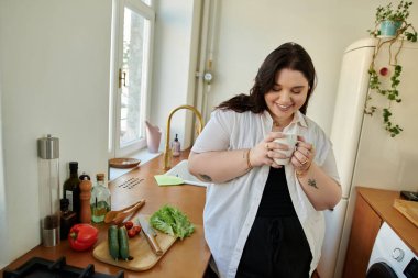 Gülümseyen kadın sıcak bir içkinin tadını çıkarıyor. Sıcak mutfağında taze malzemeler arasında..