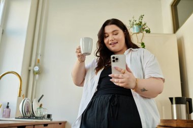 Gülümseyen kadın kahvesini yudumlar ve telefonunu mutfak ortamında kontrol eder..