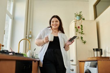 Evde dinlenirken, iri yarı bir kadın gülümserken elinde bir kupa ve bir telefon tutuyor..
