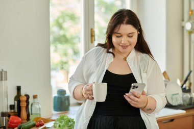 Büyük beden bir kadın, sıcak bir mutfakta telefonunu karıştırırken kahvesinin tadını çıkarır..