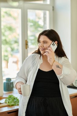 Neşeli bir kadın, elinde kahve fincanıyla telefonuyla konuşuyor..