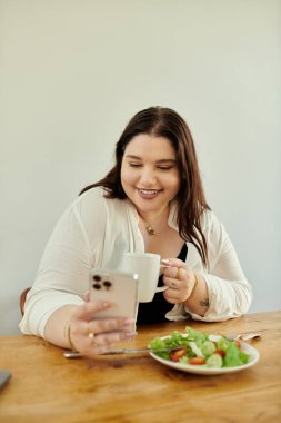 Gülümseyen kadın telefonunu kontrol ederken kahvesini yudumluyor ve sağlıklı bir yemeğin tadını çıkarıyor..