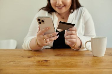 Neşeli bir kadın telefonuyla etkileşime girer, kahvesini yudumlarken alışveriş yapmaya hazırdır..