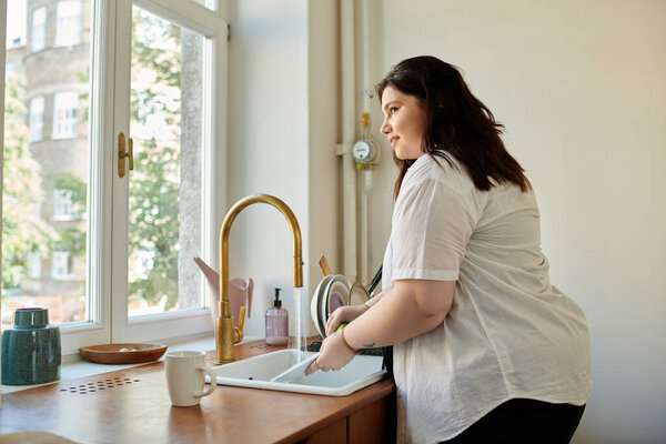 Веселая женщина плюс размер моет посуду у окна в своей уютной кухне.