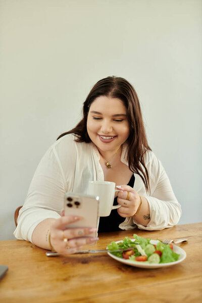 Smiling woman sips coffee while checking her phone and enjoying a healthy meal.