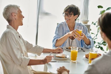 A cozy morning as lesbian parents enjoy breakfast with their adult son, filled with laughter and connection. clipart