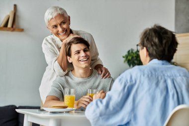 Lezbiyen ebeveynler, gülümseyen yetişkin oğullarıyla keyifli bir kahvaltıyı paylaşıyor, kahkaha ve aşktan zevk alıyorlar..