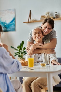 Lezbiyen ebeveynler güneşli bir sabahta gülümseme ve sevgiyle dolu yetişkin oğullarıyla sıcak bir kahvaltı yaparlar..