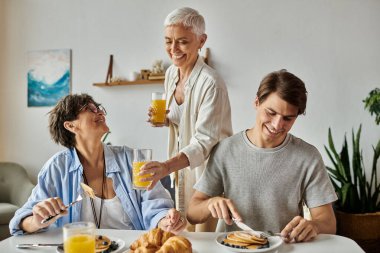 Lesbian parents joyfully share breakfast with their adult son, savoring pancakes and laughter together. clipart