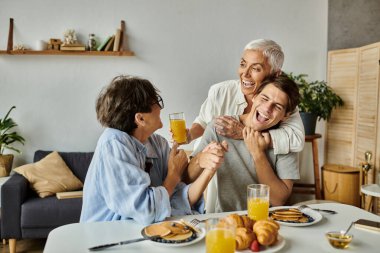 Lezbiyen ebeveynlerin ve yetişkin oğullarının kahkahalarını ve lezzetli yemeklerini paylaştığı canlı bir aile kahvaltısı..