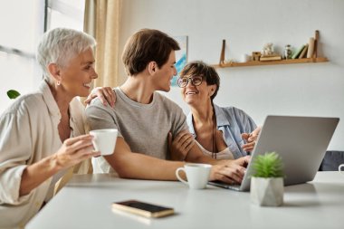 Lezbiyen ebeveynler, güneşli bir yerde kahve ve dizüstü bilgisayarla yetişkin oğullarıyla kahkahalarını ve sevgilerini paylaşıyorlar..