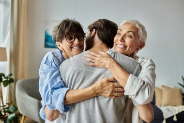 Lesbian parents and their adult son share warm hugs and laughter in their cozy living room. clipart