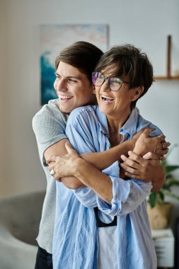 Bir anne ve yetişkin oğlu, güneşli oturma odalarında kucaklaşırken kahkahalar ve sıcaklığı paylaşırlar..