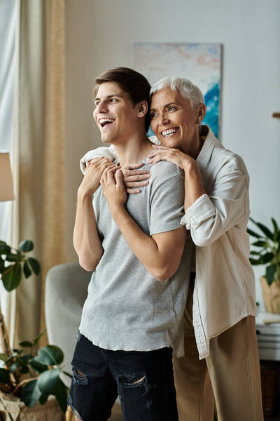 Мать средних лет и ее взрослый сын разделяют радостный момент вместе, обнимаясь в яркой гостиной.