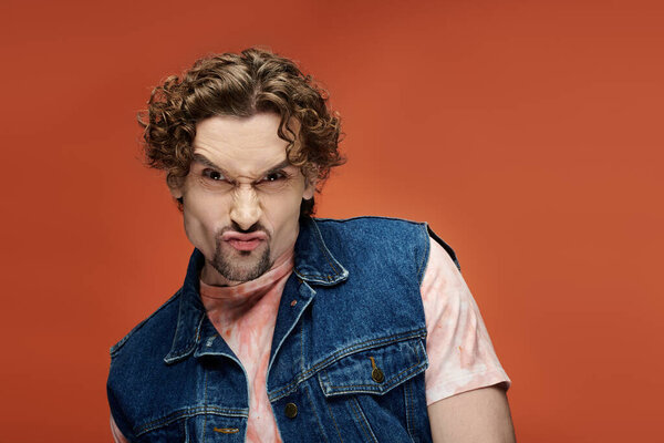 Мужчина с вьющимися волосами эмоционально позирует на ярком оранжевом фоне.