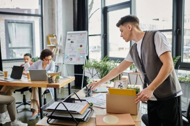 Young dapper man packing his items during lay off, colleagues on backdrop. clipart