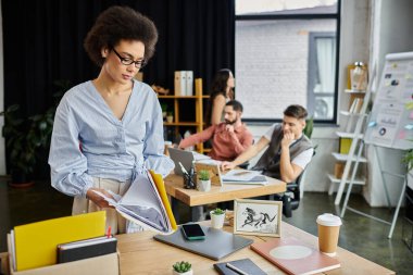Şık Afro-Amerikalı kadın işten çıkarılma sırasında eşyalarını topluyor, iş arkadaşları arka planda..