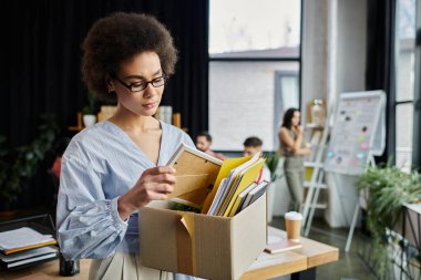 African american woman in elegant attire packing her items during lay off, colleagues on backdrop. clipart