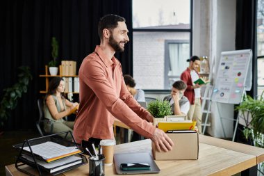 Modern ofiste işten çıkarılma sırasında eşyalarını toplayan zarif bir adam, arka plandaki meslektaşları....