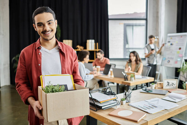 Man express emotions while packing belongings in an elegant workspace, colleagues on backdrop.