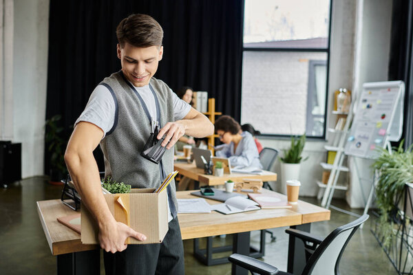 Young delighted man packing his items during lay off, colleagues on backdrop.