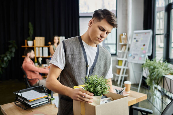 Young dedicated man packing his items during lay off, colleagues on backdrop.