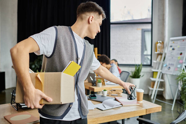 Young stylish man packing his items during lay off, colleagues on backdrop.