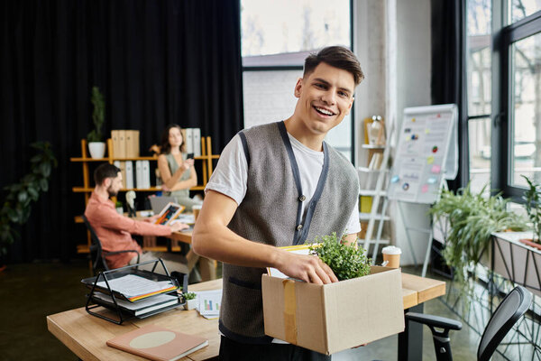 Young cheerful man packing his items during lay off, colleagues on backdrop.