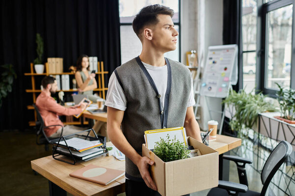 Молодой задумчивый человек собирает вещи во время увольнения, коллеги на заднем плане.