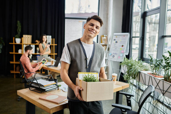 Young handsome man packing his items during lay off, colleagues on backdrop.