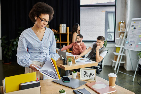 Chic african american woman packing her items during lay off, colleagues on backdrop.