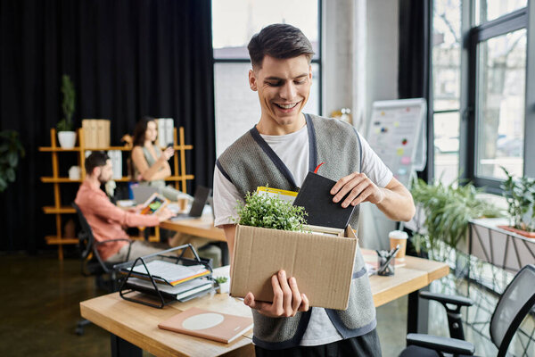 Young jolly man packing his items during lay off, colleagues on backdrop.